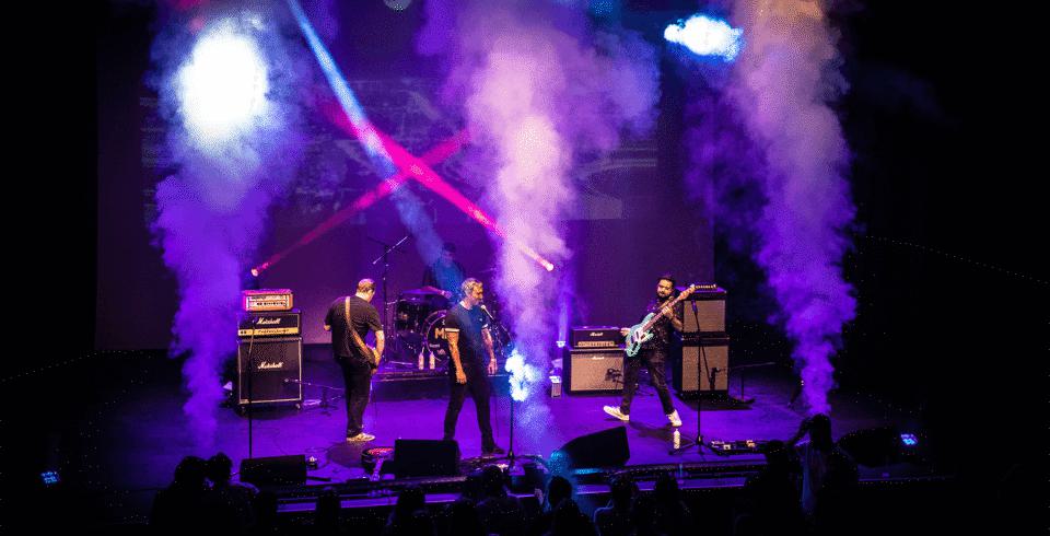 Surrounded by upward jets of fog and lit by blue light a band perform on stage