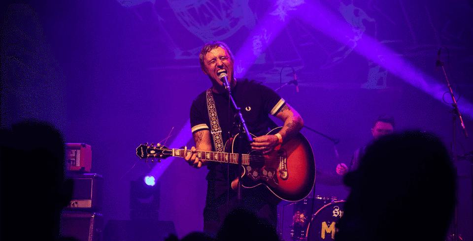 Lit by blue light and wearing a black t-shirt a man sings while playing guitar