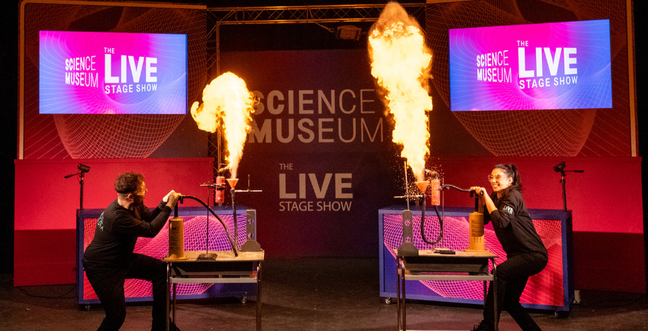 One man and one woman completing a science experiment on stage with large flames. the background is a pink and purple colour.