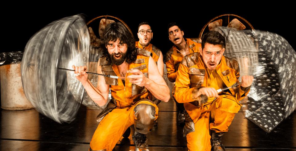 Four men dressed in yellow jumpsuits twirl clear plastic umbrellas in unison