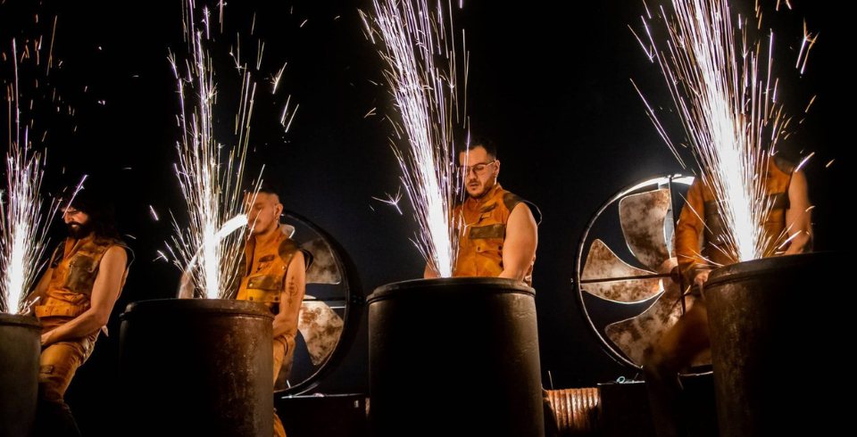 Sparks fly on metal barrels while four men stand behind them in yellow suits on stage