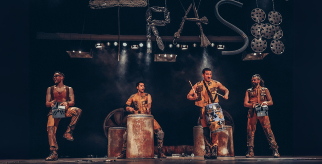 Four men drum on a variety of metal tins and drums while the word Trash is suspended above them, made out of scrap metal