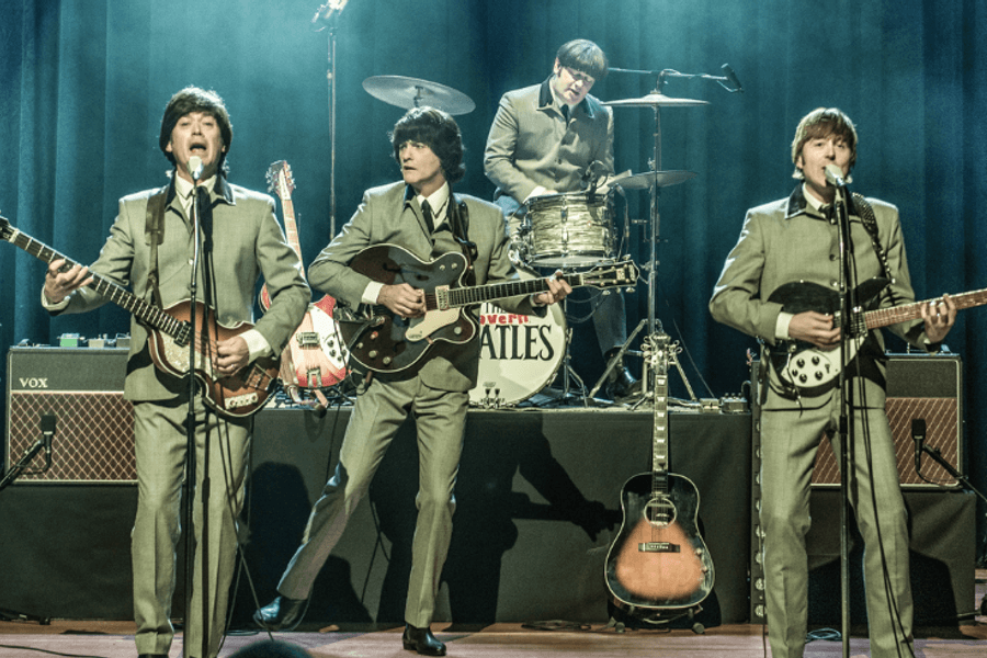 four men dressed in dark grey suits performing on stage. all have dark brown/black bobbed hair.