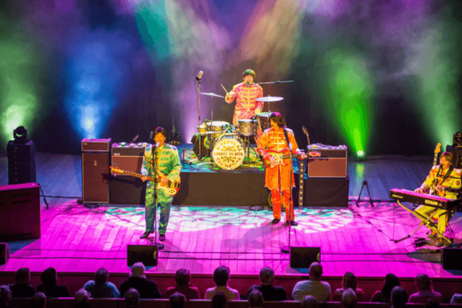 A group of performers on stage in a band with multi-coloured outfits under pink lighting