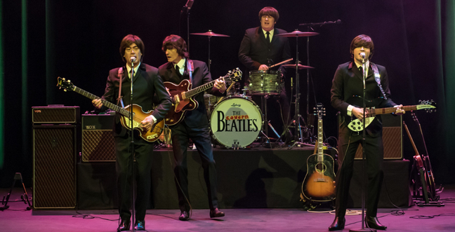 four men dressed in dark grey suits performing on stage. all have dark brown/black bobbed hair.