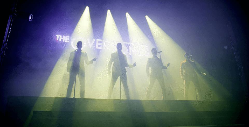 Four men stand in the yellow spotlight on stage with their silhouettes
