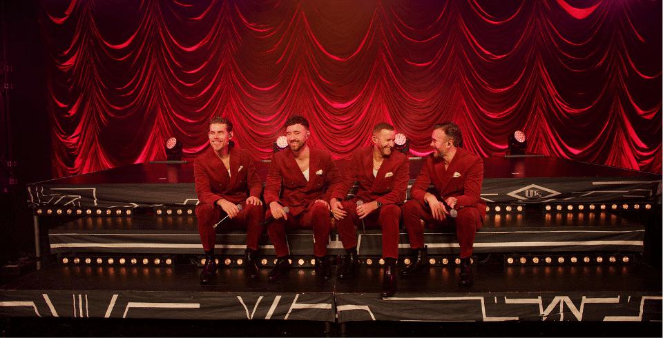 Four men sit on stage in a red suits ribbed red curtain behind them