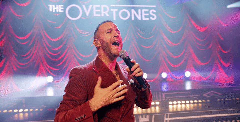 A man sings in a red suit with a ribbed red curtain behind him and the words 'The Overtones' lit up on stage