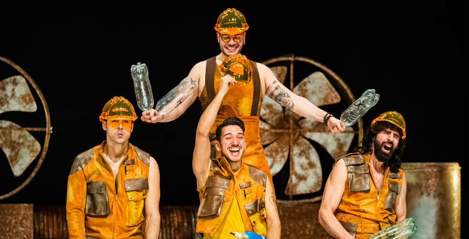 Three men sit on the floor wearing yellow hardhats while a fourth man stands behind them and hits them on the head with plastic bottles