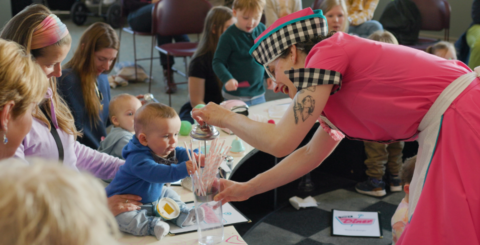 A group of children around a table with a woman dressed as a diner lady while she plays with a baby and straws