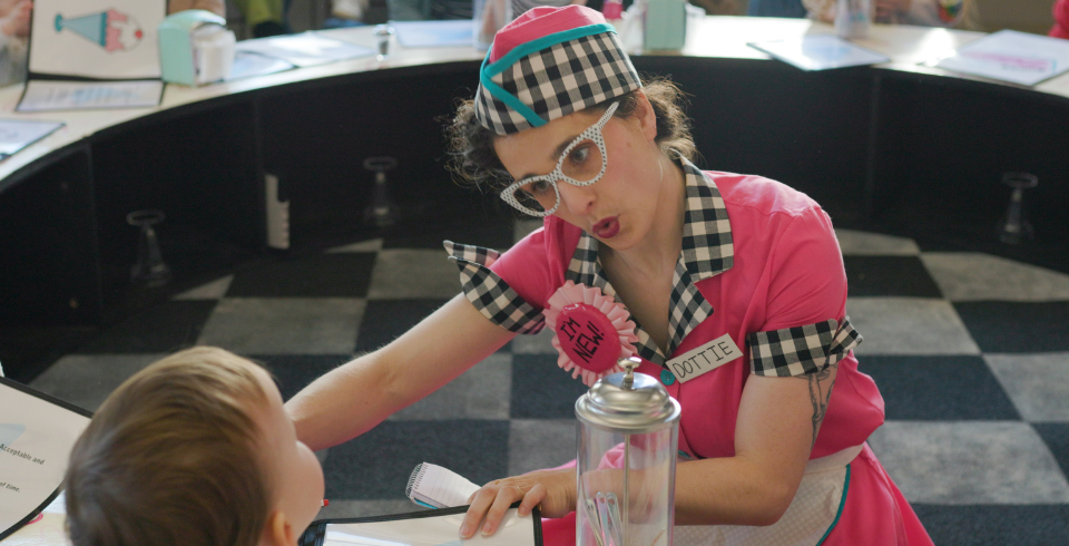 A woman dressed as a diner lady in pink with black and white check sits at a table with a child