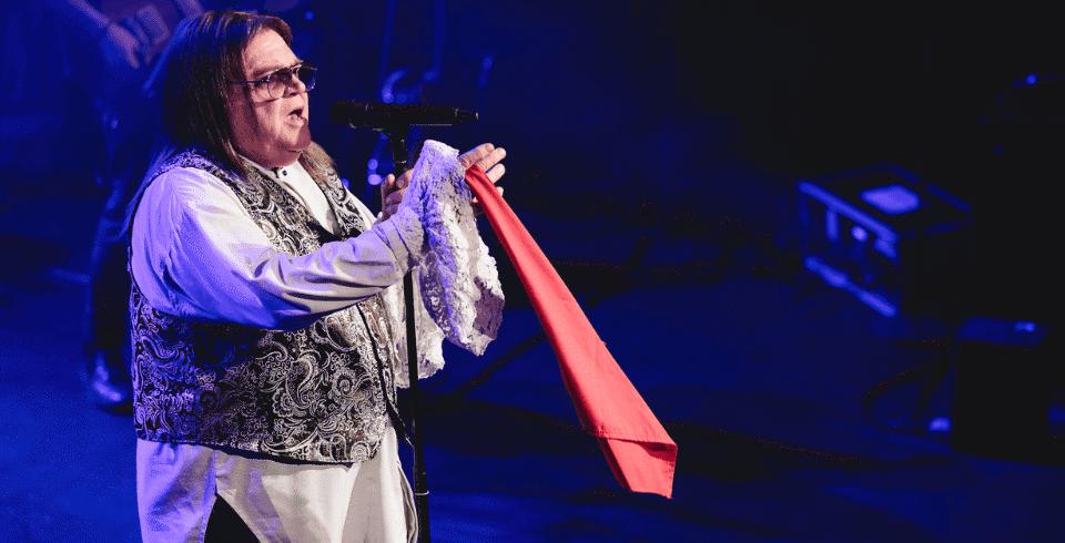 A man with long brown hair, a white shirt and a silver waistcoat singing into a microphone with a red scarf in his hand