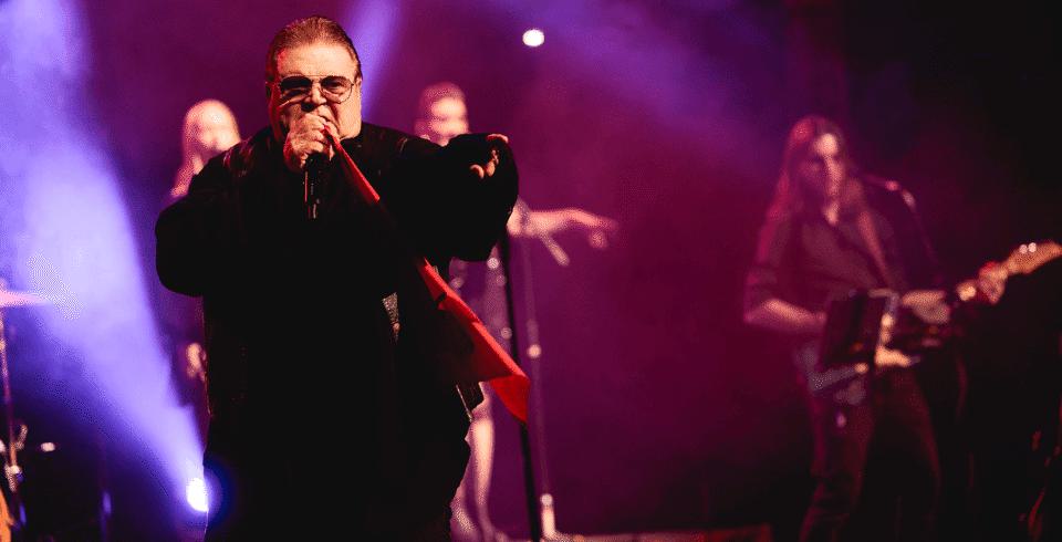A man in a black top on stage singing into a microphone in front of purple back lights