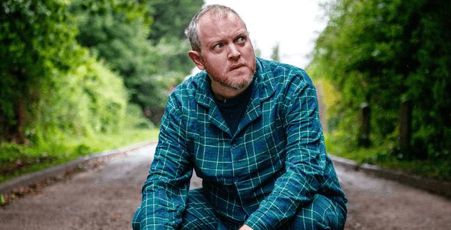 A white man with short hair crouches on the ground in the countryside wearing green plaid pyjamas