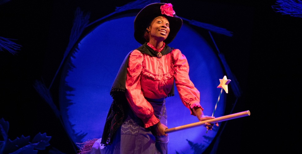 A woman with a red flower on her hat wears a red shirt while holding a star and on a broom