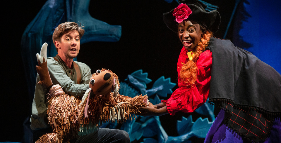 A woman with a red flower on her hat shakes hands with a shaggy brown dog puppet on stage