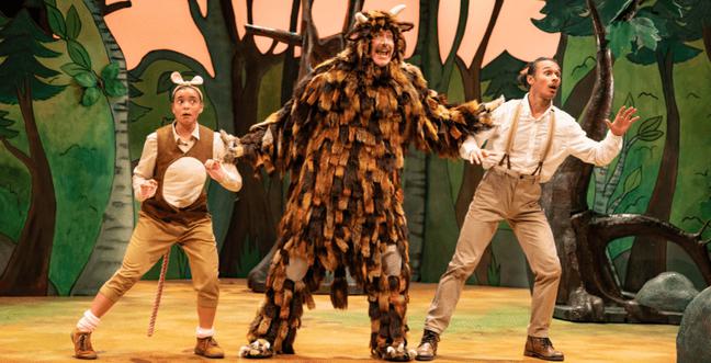 Three people dressed as woodland creatures in beige and brown trousers and waistcoats on stage, with one dressed as a Gruffalo against a green forest