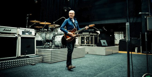 A man holding an electric guitar stands in front of white band equipment