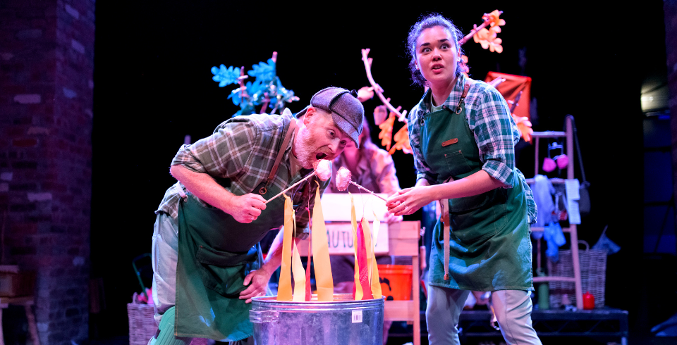 Two people on stage in green overalls and check green shirts stand over a fake fire made out of paper with marshmallows on sticks