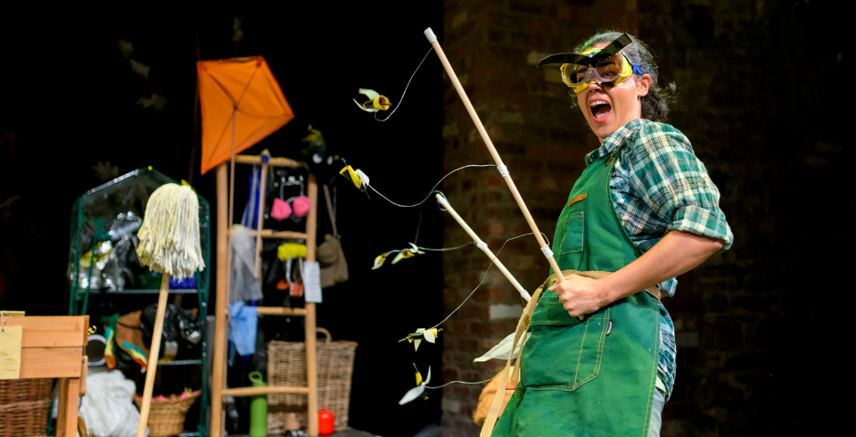 A woman stands in green overalls and a check shirt holds a kite while wearing goggles