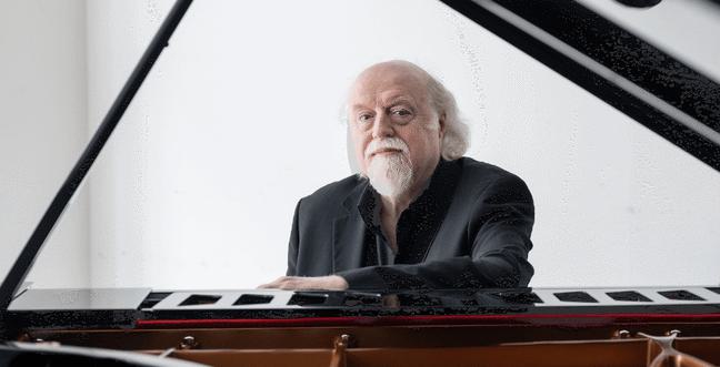 An older man with white hair and a beard sits at a grand piano, wearing a black suit, with a calm expression.
