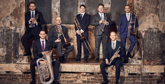 A group of men in suits stood on a historical building backdrop. Each man is holding a brass instrument