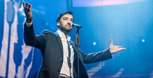 A man in a suit stands in front of a microphone on a blue lit stage with his arms out