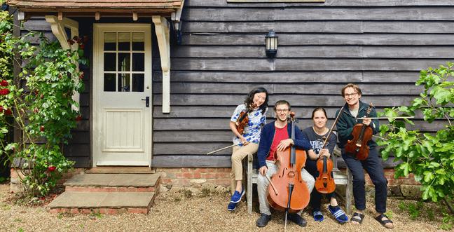 Four people sat on a bench outside a building, each holding a different string instrument including cello and violin