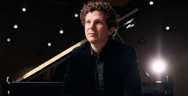 Lit from behind a man dressed in black sits with his back against a grand piano