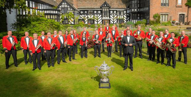 A large group of musicians dressed in black trousers, white shirts and red blazers stand in a green grassy courtyard space