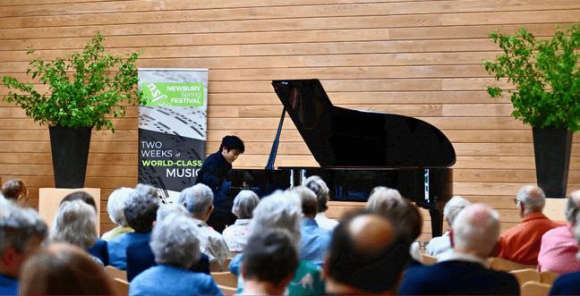 A pianist sits at a grand piano in front of a an audience of people in a wooden room with large green plants in black plant pots beside the piano