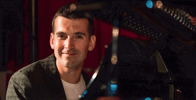 A man in a blazer sits at a grand piano in low light