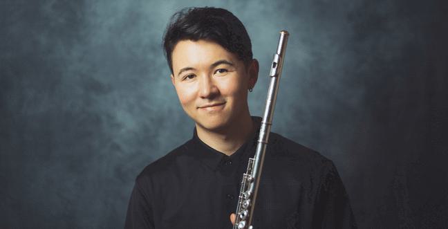 A man with black hair and wearing a black shirt stands in front of a grey backdrop holding a flute