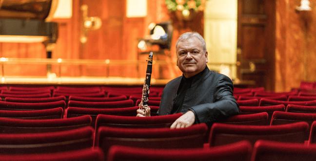 A man sat in red chairs by a stage wears a black suit and holds a flute