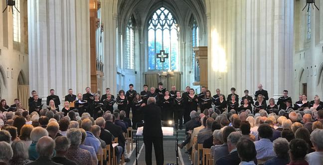 A choir stand at the front of a church with a conductor standing in the middle of the aisle and a crowd of people sat watching
