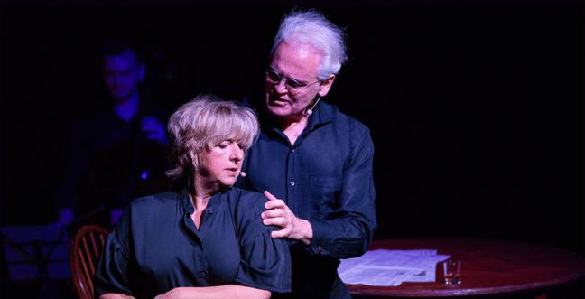 A man and a woman on stage wearing black shirts. The man has white hair and holds the woman's shoulder.