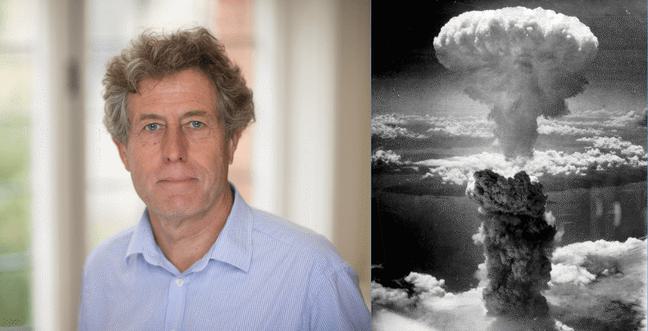 A white man with grey hair wears a blue shirt standing in front of a window. Next to him is an image of a nuclear bomb explosion in black and white