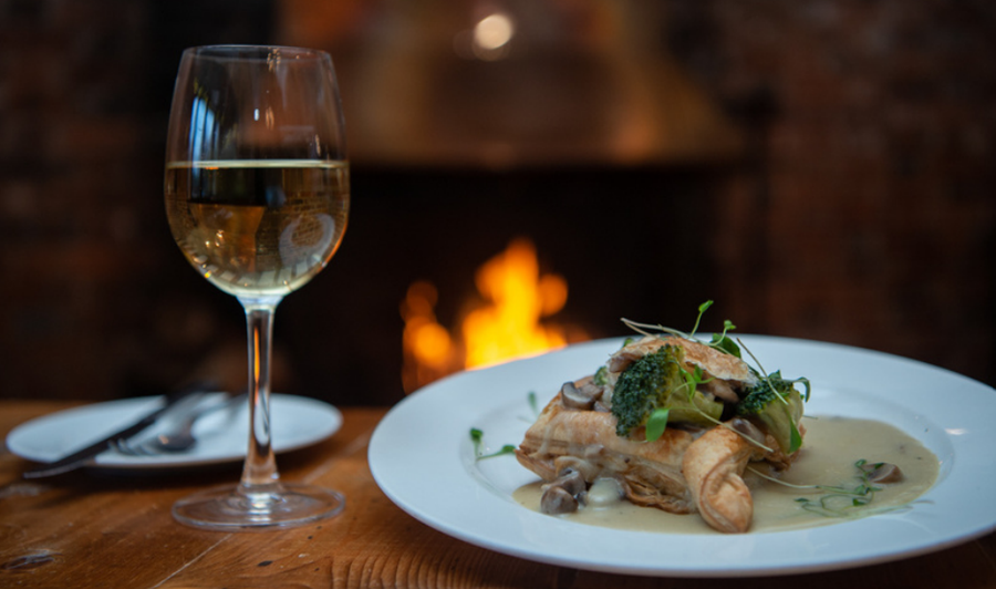 A glass of white wine and a meal on a white plate sits on a wooden table