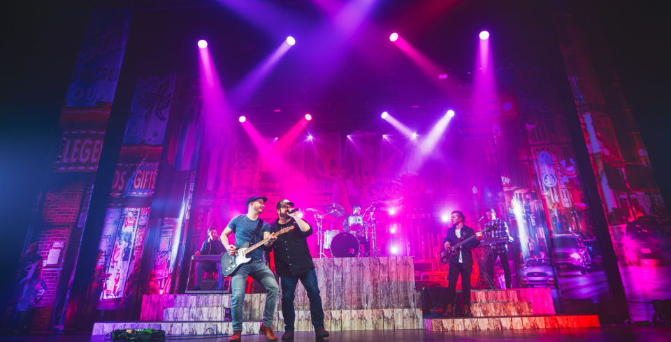 A country musician performs on stage with a guitar, surrounded by a band and illuminated by vibrant pink and purple lights.