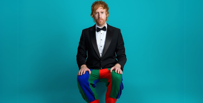 A man in a tuxedo with colorful patchwork trousers sits against a gray background, wearing a serious expression.