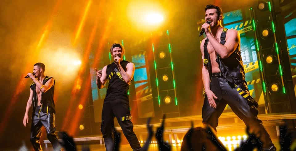 three men on stage in front of a glowing yellow light wearing leather black waistcoats and black trousers