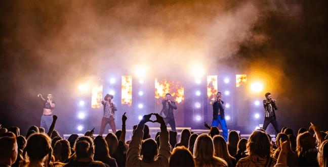 A boy band group on a glowing yellow stage with a crowd, in the middle a person holds their hands up in a heart