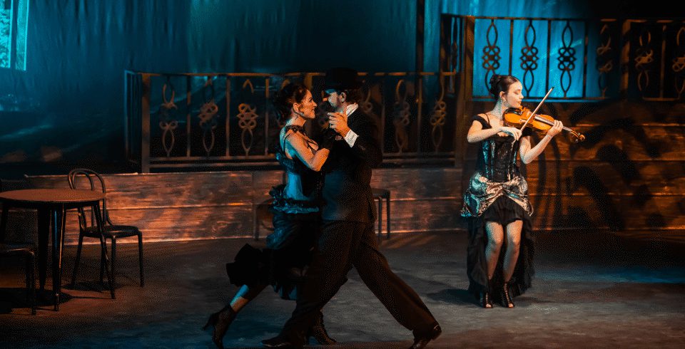 A couple dance on stage with a woman on a violin in the background