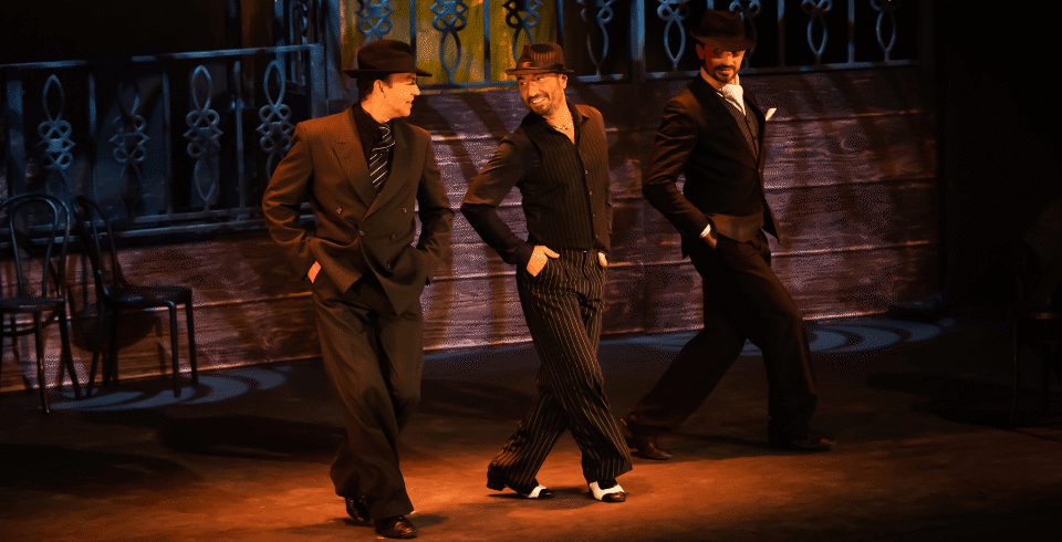 Three men in black shirts and trousers wear hats. They walk together on a dark stage, wearing tap shoes