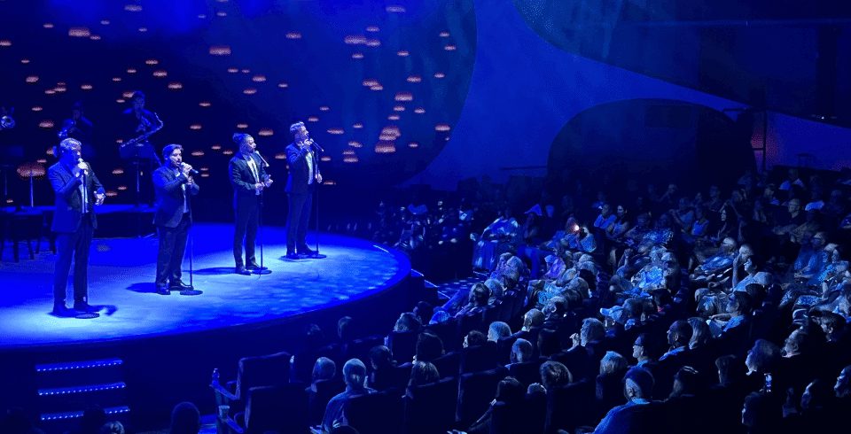 A group of people singing into microphones on stage in suits under blue lights and a large crowd