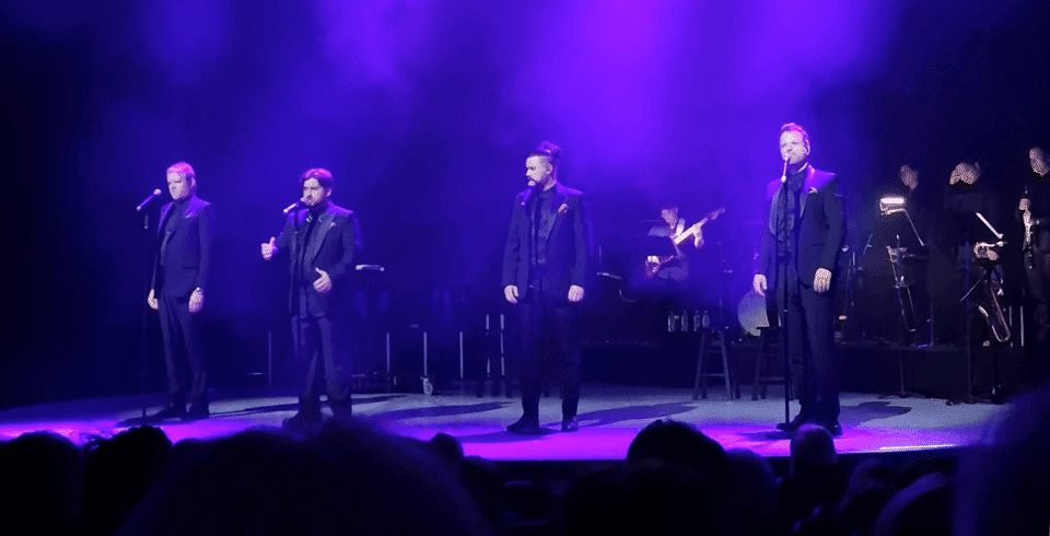 A group of people singing on stage in suits under purple lights