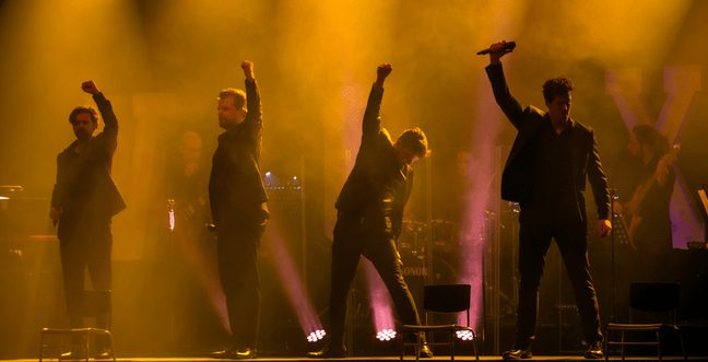 Four men in black shirts and blazers, raising their hands up on stage, in front of orange spotlights. One man has a microphone.