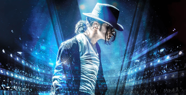 Man stood in blue lighting looking down. With long dark hair in a ponytail wearing a black hat and shirt and jacket