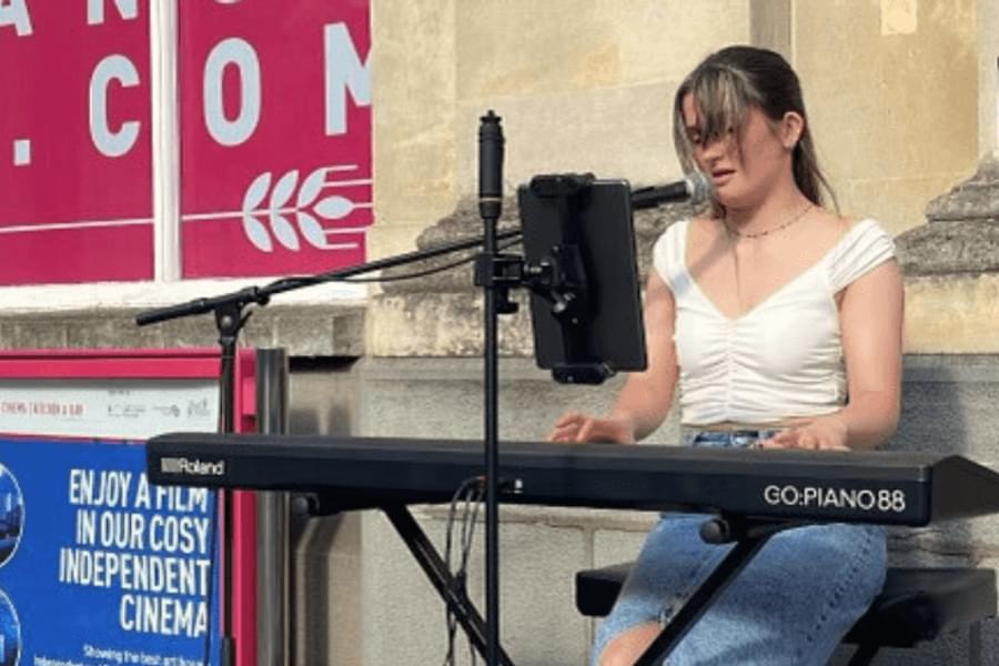 A girl in a white top and denim midi skirt sits at a keyboard outside the Corn Exchange