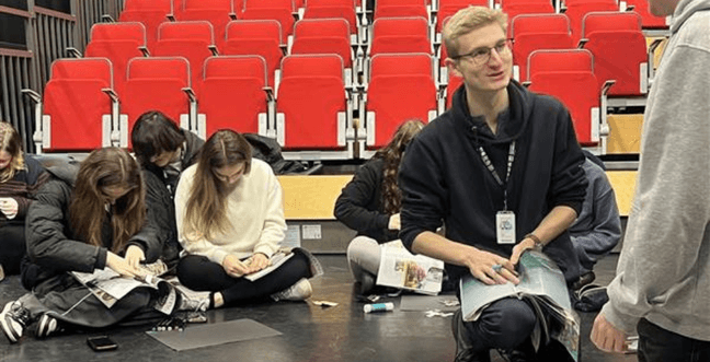 A group of people sit cross-legged on the floor while cutting out paper in front of rows of red seats. A man in glasses wearing a lanyard leans on his kneed talking to someone